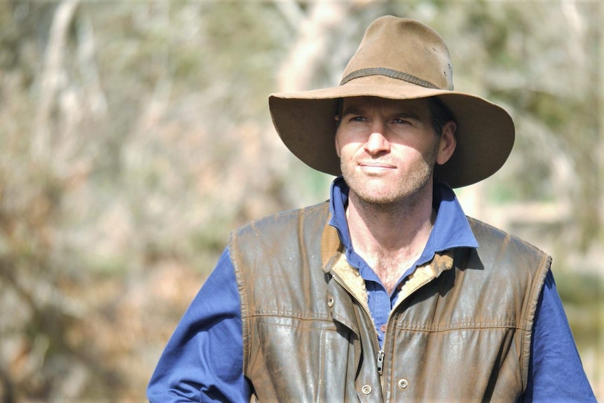 A man wearing a hat and a leather waistcoat stands in front of rural scenery.