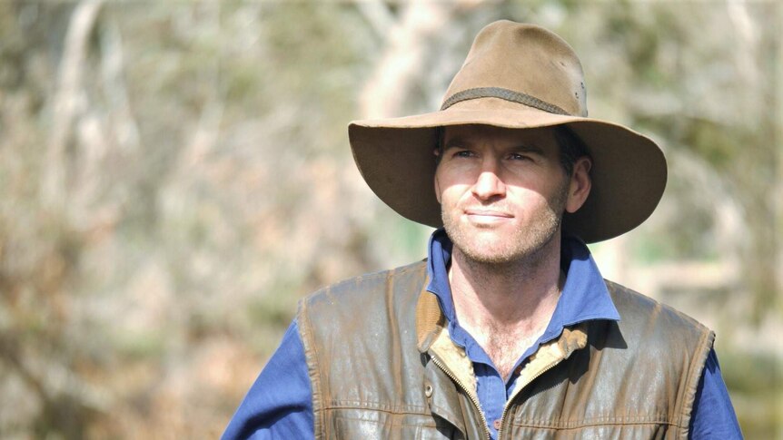 A man wearing a hat and a leather waistcoat stands in front of rural scenery.