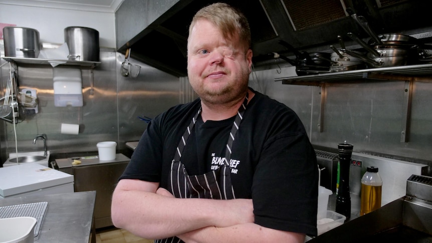 A man who has his left eye removed smiles while wearing a chefs apron, standing inside a commercial kitchen