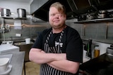 A man who has his left eye removed smiles while wearing a chefs apron, standing inside a commercial kitchen