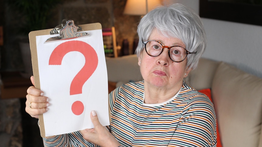 A grey haired woman holding up a clipboard with a red question mark on it