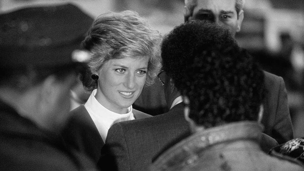 Black and white photo of Princess Diana with short blonde hair smiling through gritted teeth and surrounded by press.