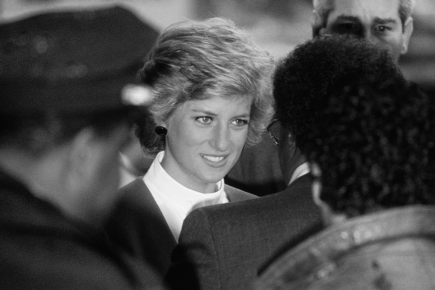 Black and white photo of Princess Diana with short blonde hair smiling through gritted teeth and surrounded by press.