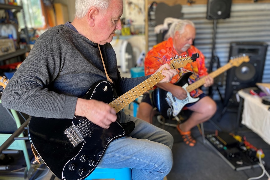Two older men sit next to each other playing guitars.
