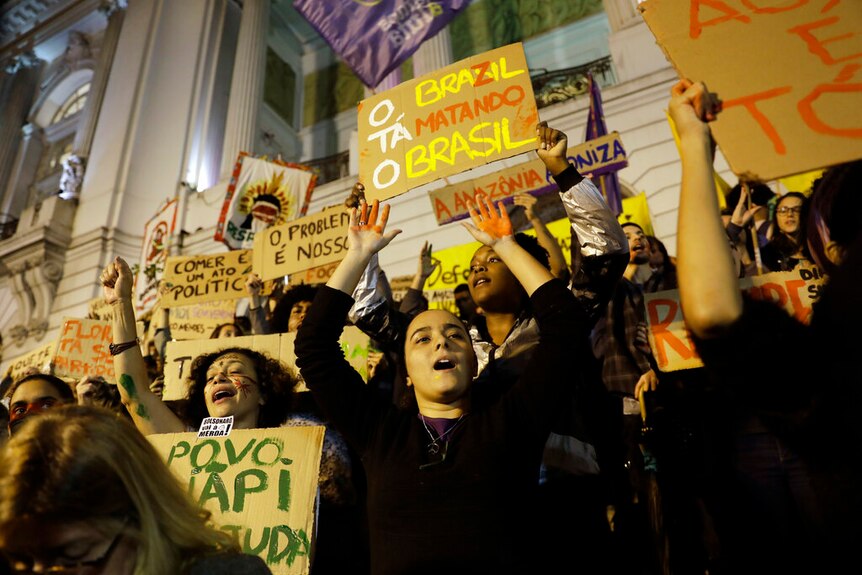 A crowd of protesters holding placards above their heads, protest outside a a building in Rio de Janeiro.
