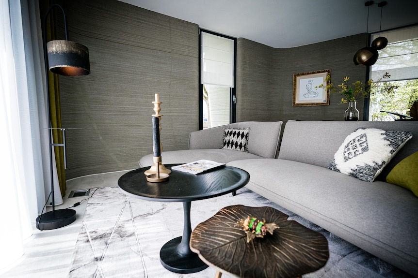 An interior of a small home shows a grey couch, a coffee table and some lamps.