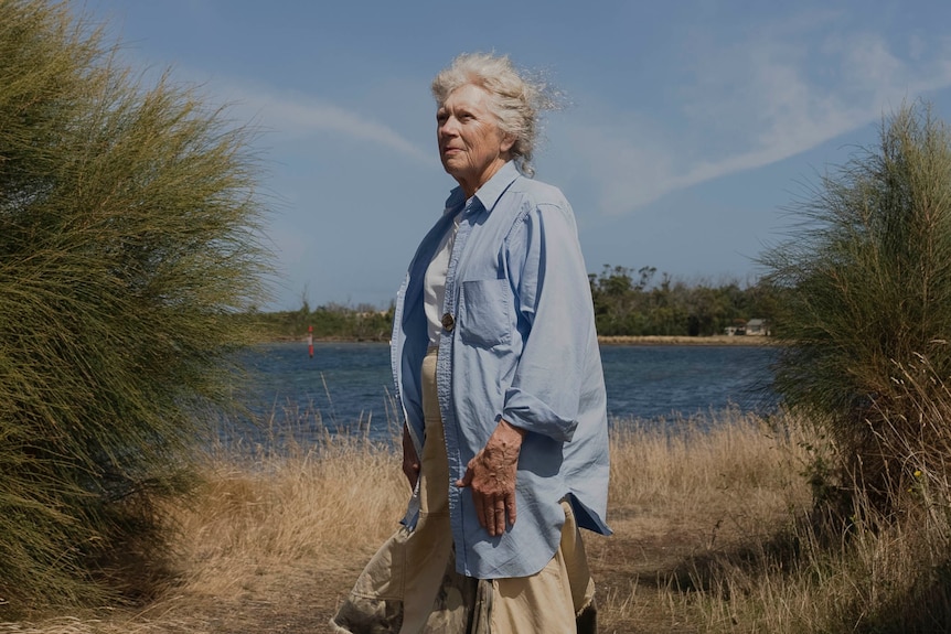 a woman in a blue loose shirt and a skirt standing near the water, hair blowing in the wind