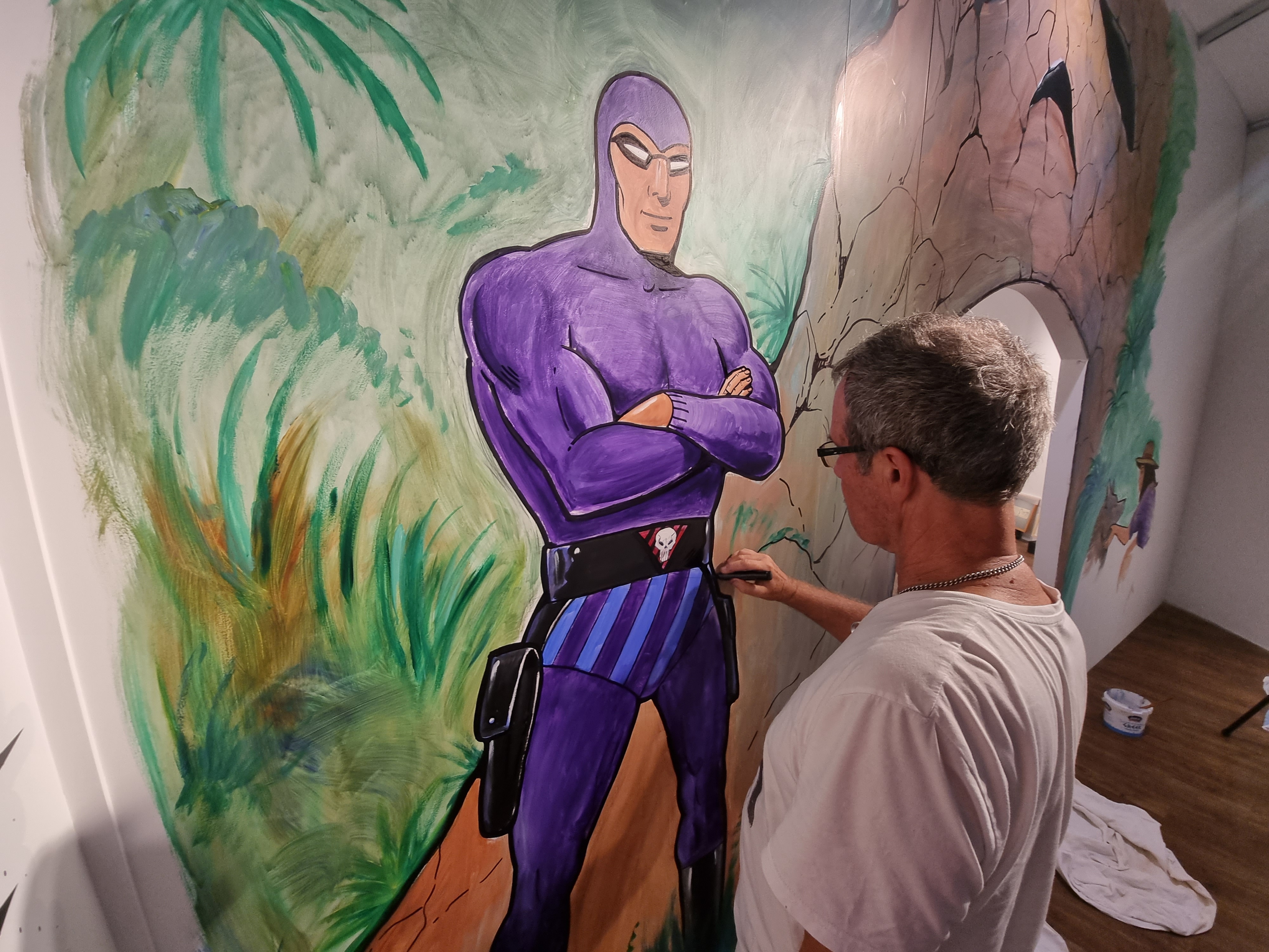 A man painting a mural on a large wall. The mural is a giant purple superhero, The Phantom