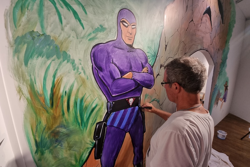 A man painting a mural on a large wall. The mural is a giant purple superhero, The Phantom