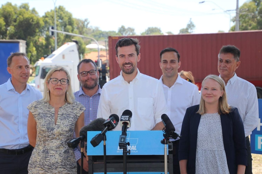 A wide shot with Zak Kirkup standing centred at a podium with microphones, with other people surrounding him and a truck behind.