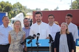 A wide shot with Zak Kirkup standing centred at a podium with microphones, with other people surrounding him and a truck behind.