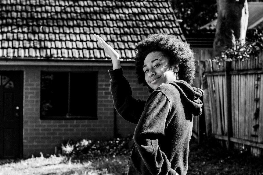 Lou is posing in her backyard in a casual friendly way. One arm on her waist, the other in the air. Afro out.