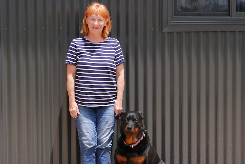 A woman with red hair stands in front of a corrugated-iron wall with a black dog.