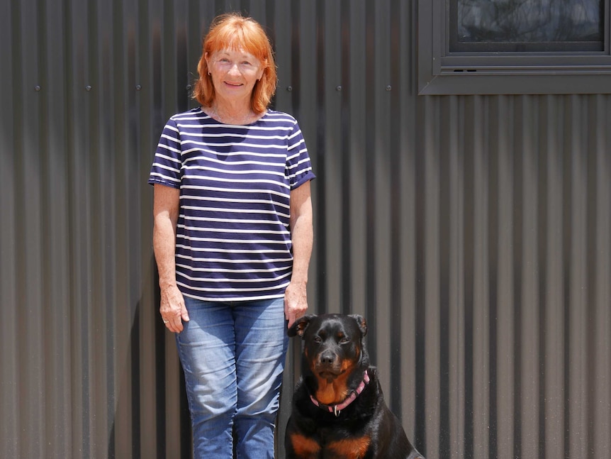 A woman with red hair stands in front of a corrugated-iron wall with a black dog.