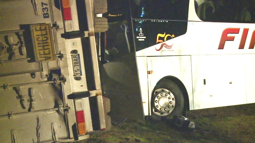 A white bus smashed into the underside of a truck trailer resting on its left-hand side on the highway.