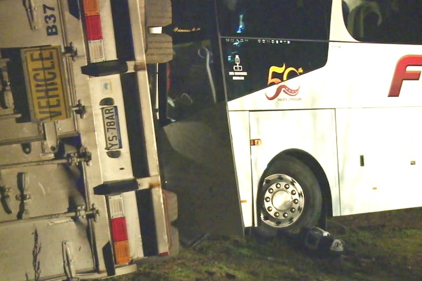 A white bus smashed into the underside of a truck trailer resting on its left-hand side on the highway.