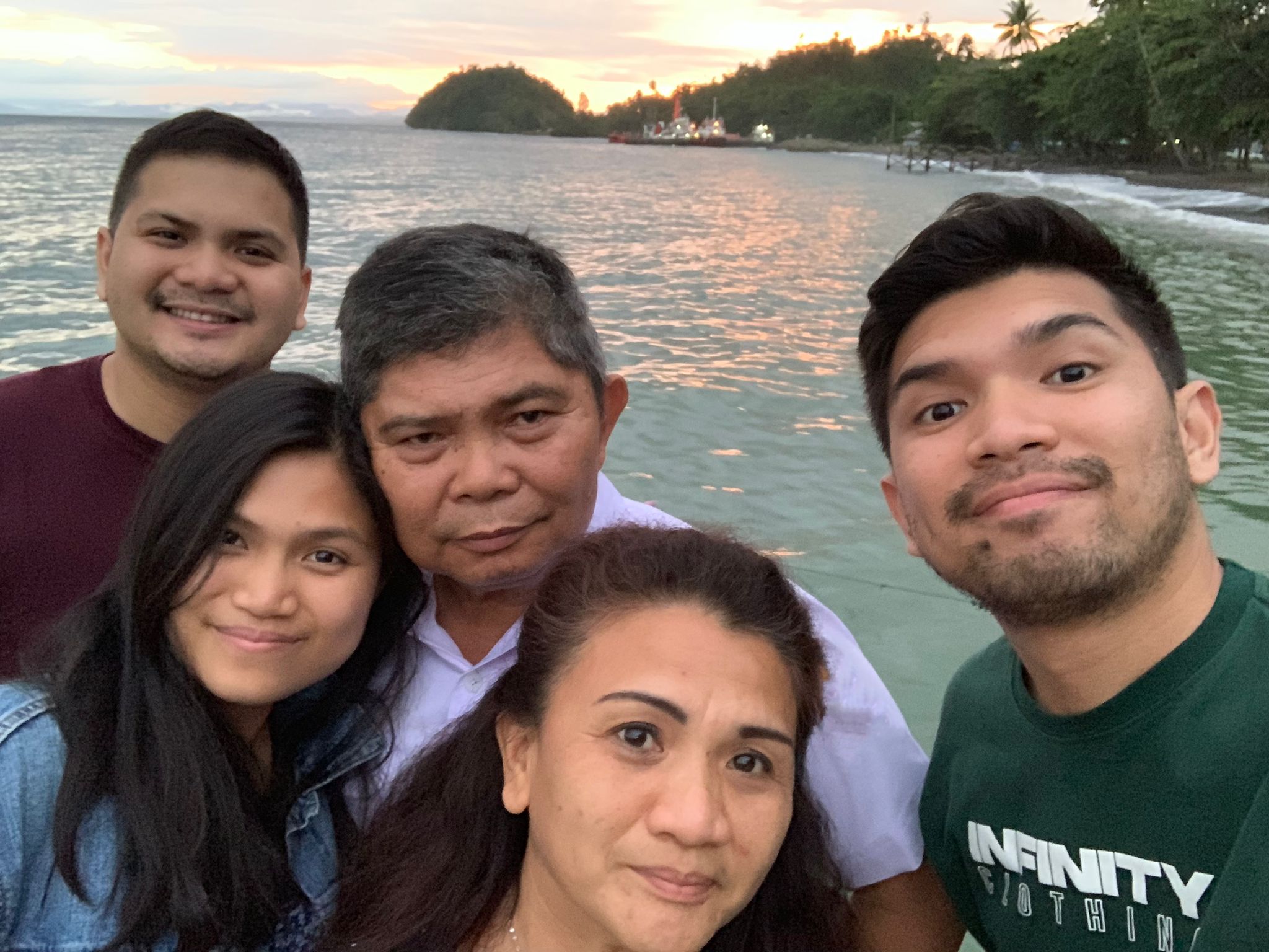 A family picture of five selfie. Parents with their adult two sons a daughter with a sea in the background.