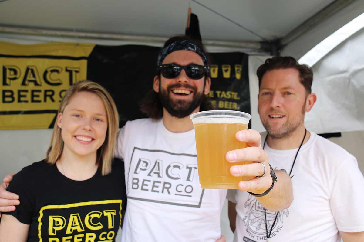 Three people, one of whom is holding a beer, stand behind portable beers taps in a festival tent.