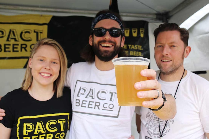 Three people, one of whom is holding a beer, stand behind portable beers taps in a festival tent.