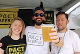 Three people, one of whom is holding a beer, stand behind portable beers taps in a festival tent.