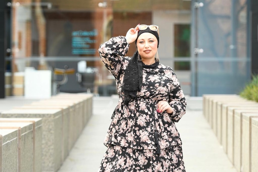 A photo of AIsha wearing hijab, in a floral dress, and wearing sunglasses over her scarf.