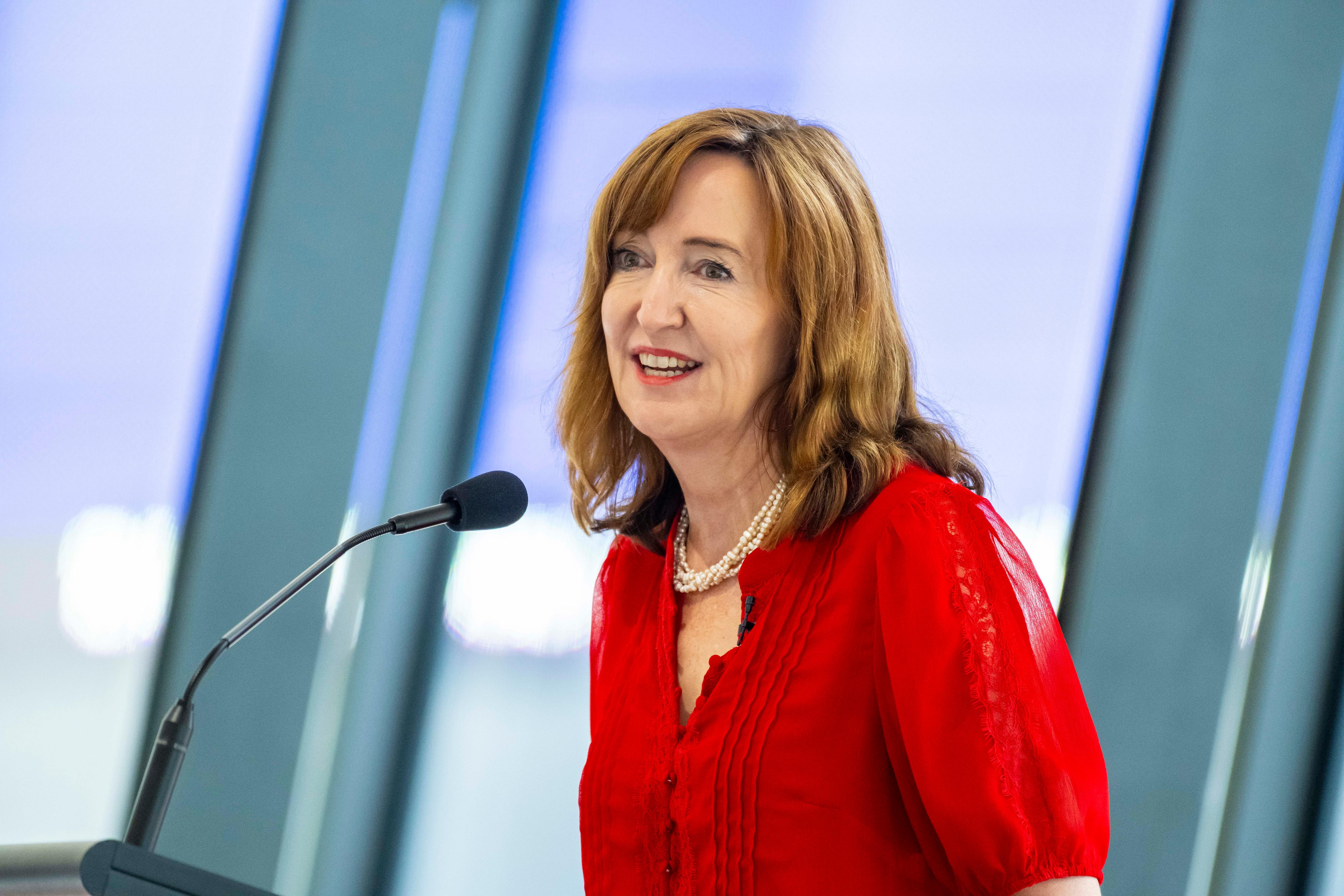 Susan Close in a red top speaks into a microphone in front of large glass windows