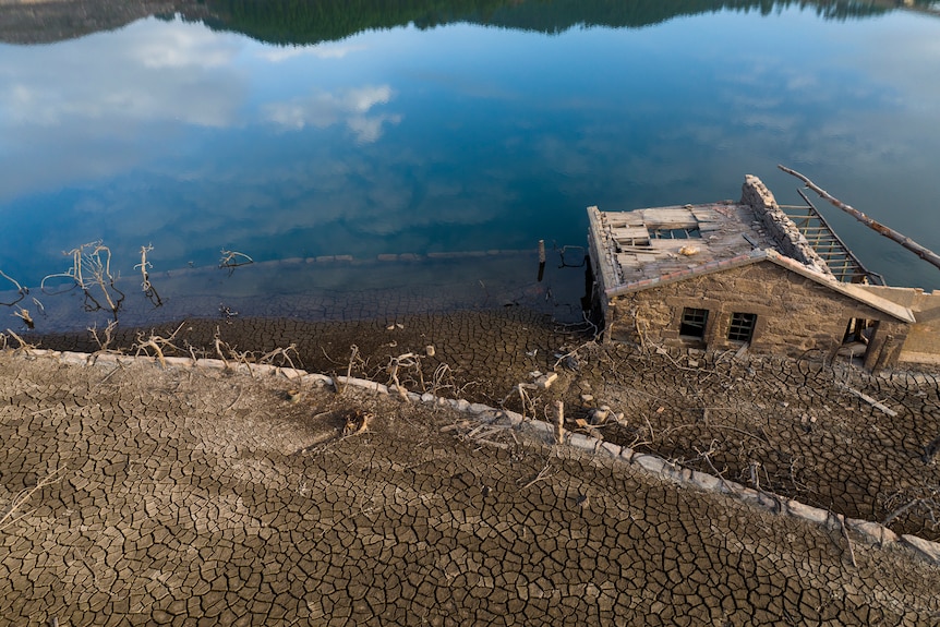 A house sits between the cracked dry Earth and the blue water of the reservoir