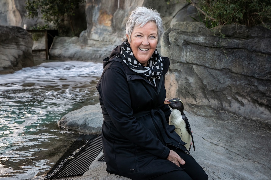 A woman with short grey hair sits next to a waterfall with a penguin next to her
