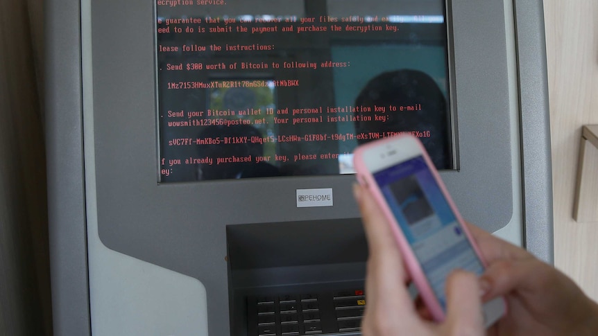 A message demanding money is seen on an ATM monitor as someone uses their iPhone in the foreground