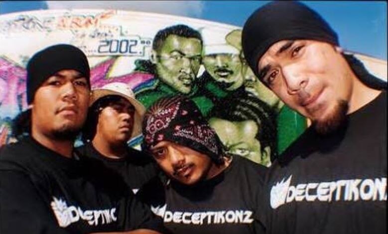 Four men wearing black teeshirts saying Deceptikonz stand infront of a colourful mural.