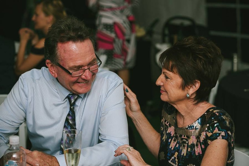 John Jeffreys and wife Yvonne Prole at a function smiling at each other, with a glass of champagne on the table in front of them