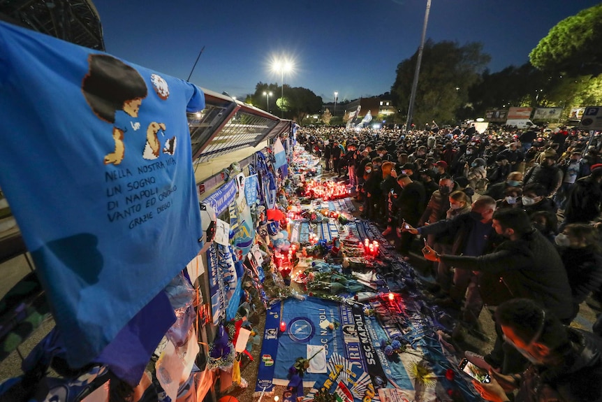 Football fans gather in front of Diego Maradona memorial filled with candles, banners and scarves.