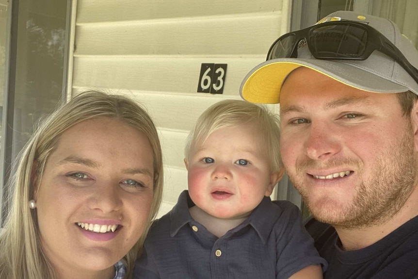 A selfie-style shot of a blonde woman and a man in a cap holding a young child.