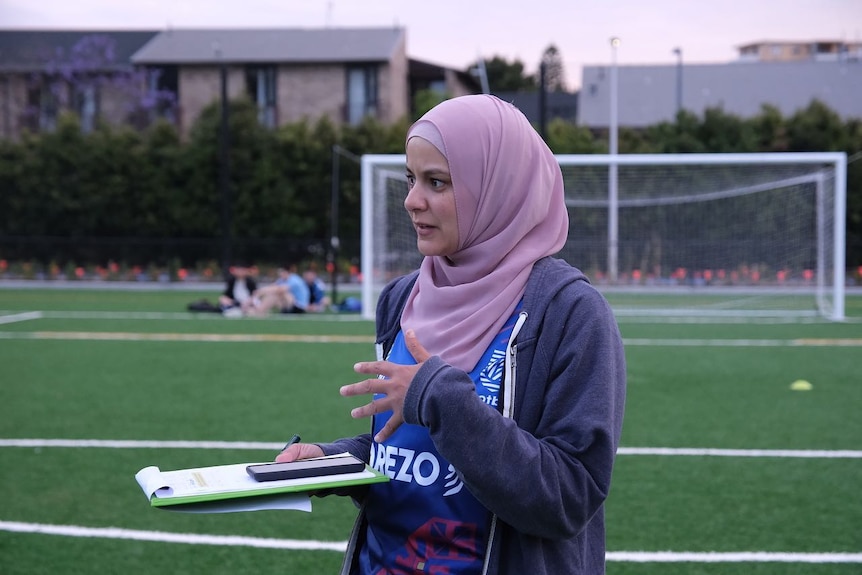 Assmaah Helal speaks while holding a clip board on a soccer field.