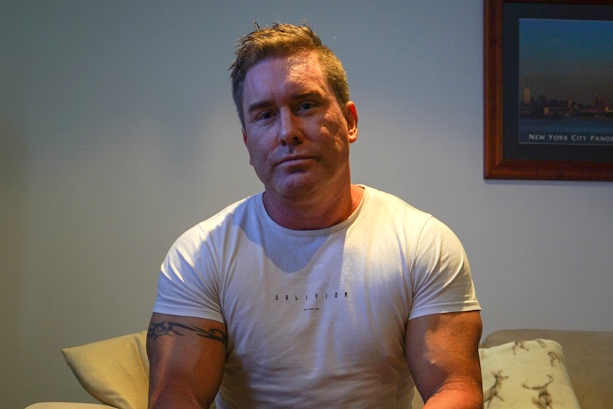 A muscular man wearing a white shirt sits on a couch.