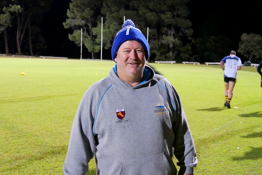 A man wearing a beanie standing in the middle of a football oval. 