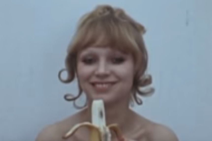 A blonde woman with bare shoulders smiles as she holds a banana.