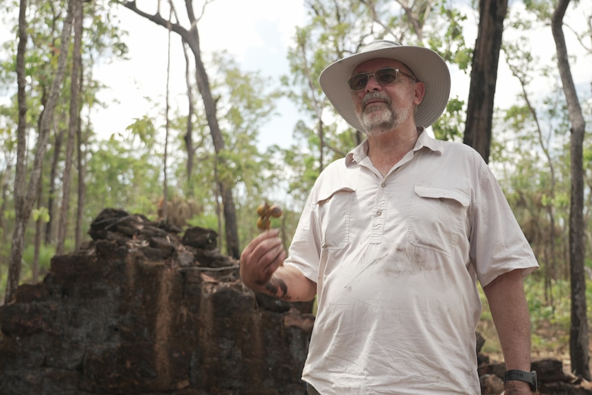An elderly Caucasian man standing next to remnants of an old brick building in a forest.