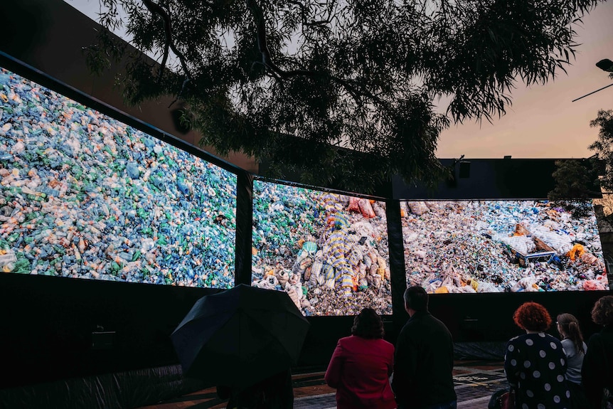 People watch screens with photographs of piles of plastic bottles in the evening