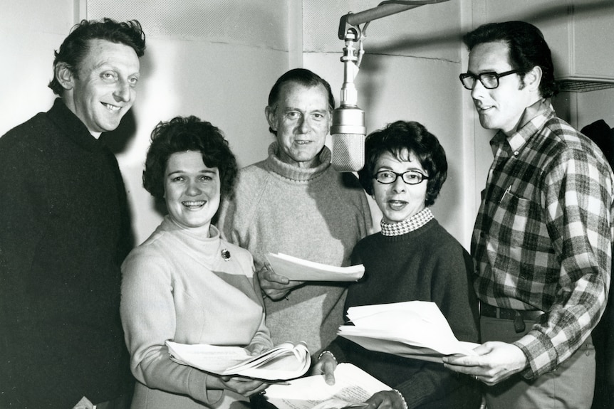A black and white image of five men and women around a microphone holding pieces of paper. 