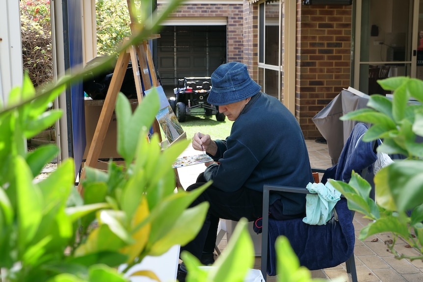 A man paints at an easel framed by trees