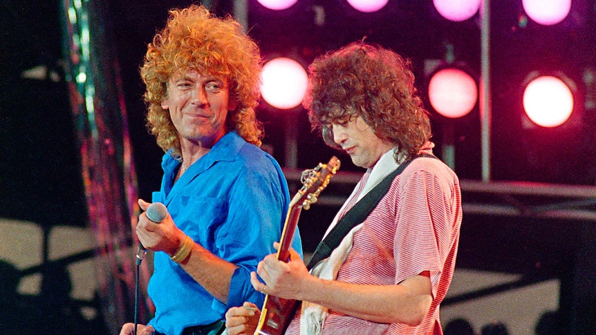 Photo from 1985 showing Robert Plant holding a microphone loosely, and looking at Jimmy Page playing a guitar intently