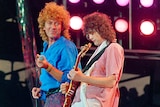Photo from 1985 showing Robert Plant holding a microphone loosely, and looking at Jimmy Page playing a guitar intently