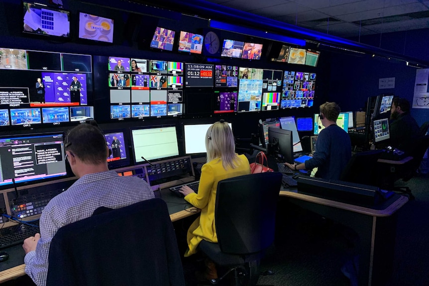 Sala de control con muchos monitores de televisión y cuatro personas sentadas en escritorios con auriculares.