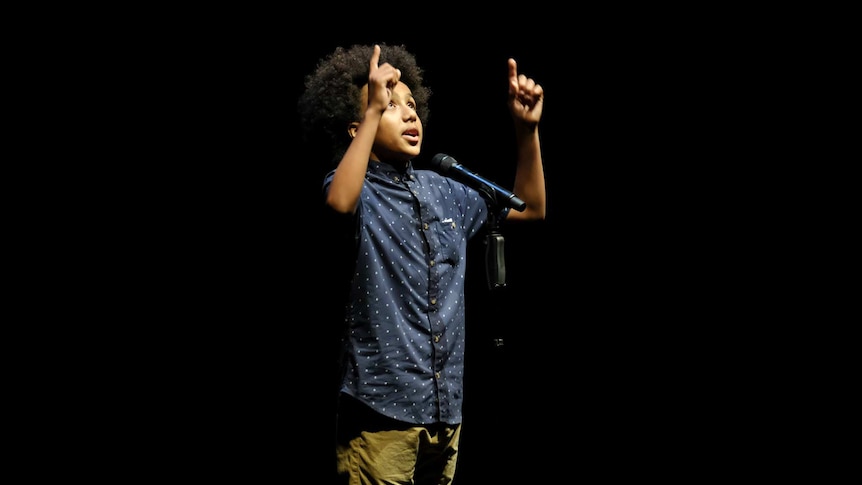 Solli pointing upwards whilst perfuming his poem. Dark lit stage.