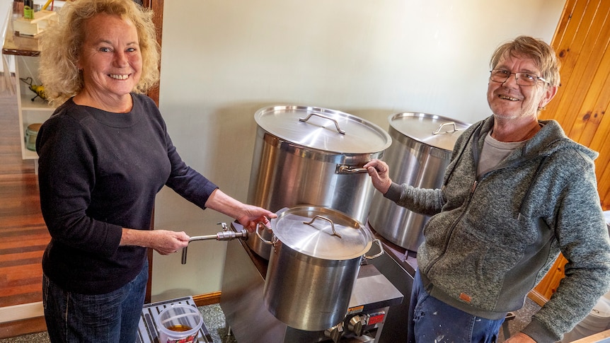 A woman and man stand beside three large cooking pots.