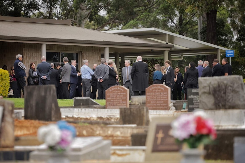 A group of people in suits talk in small groups on lawn with tombstones in the foreground.