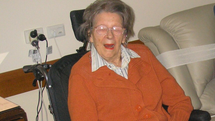 An elderly woman in an orange cardigan sits in an armchair