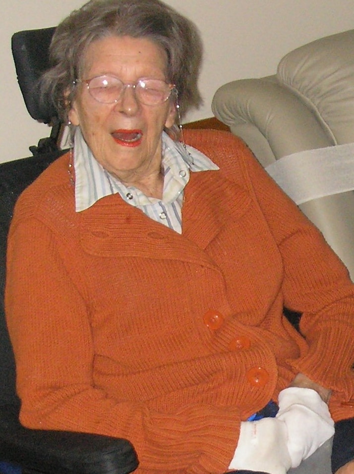 An elderly woman in an orange cardigan sits in an armchair