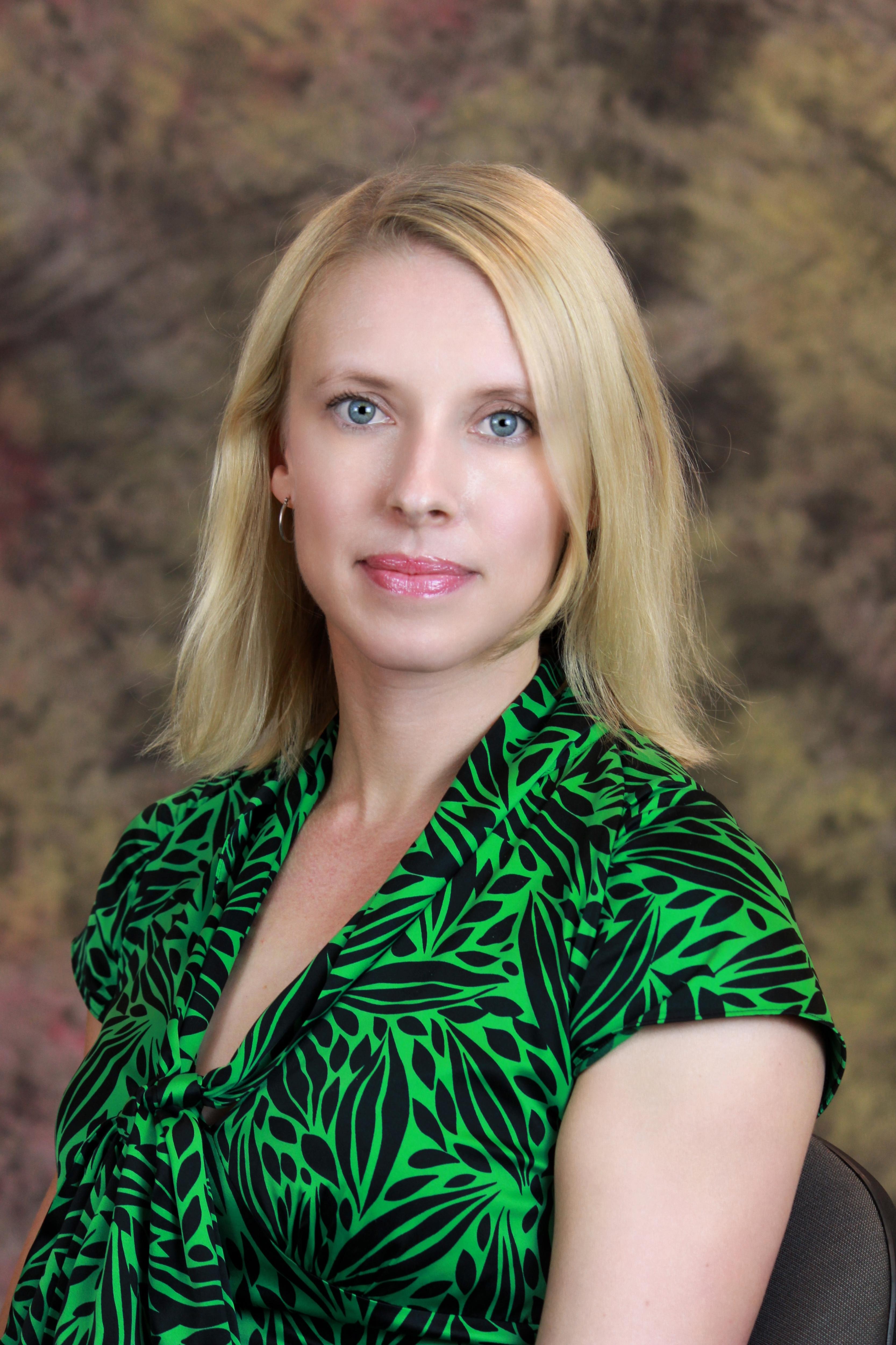 A portrait of a woman with blonde hair wearing a green shirt in front of a brown background.